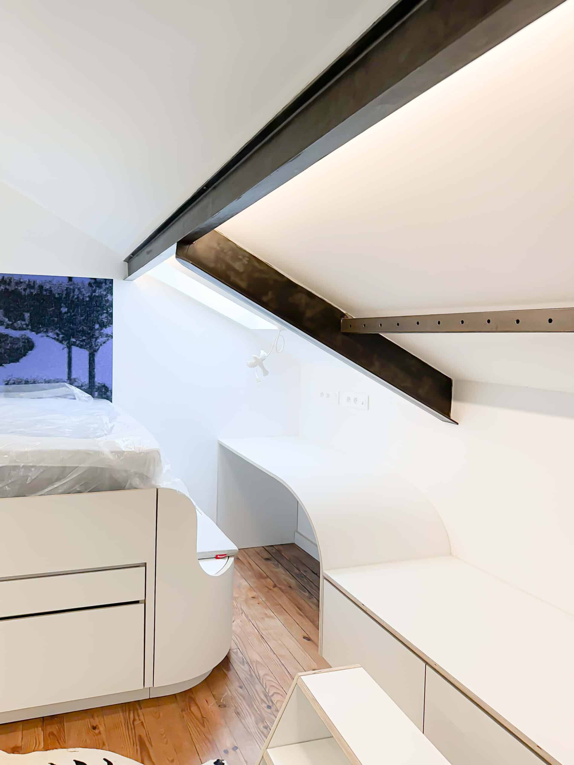 Chambre mansardée moderne, bureau blanc courbé et poutres métalliques apparentes sur parquet clair.