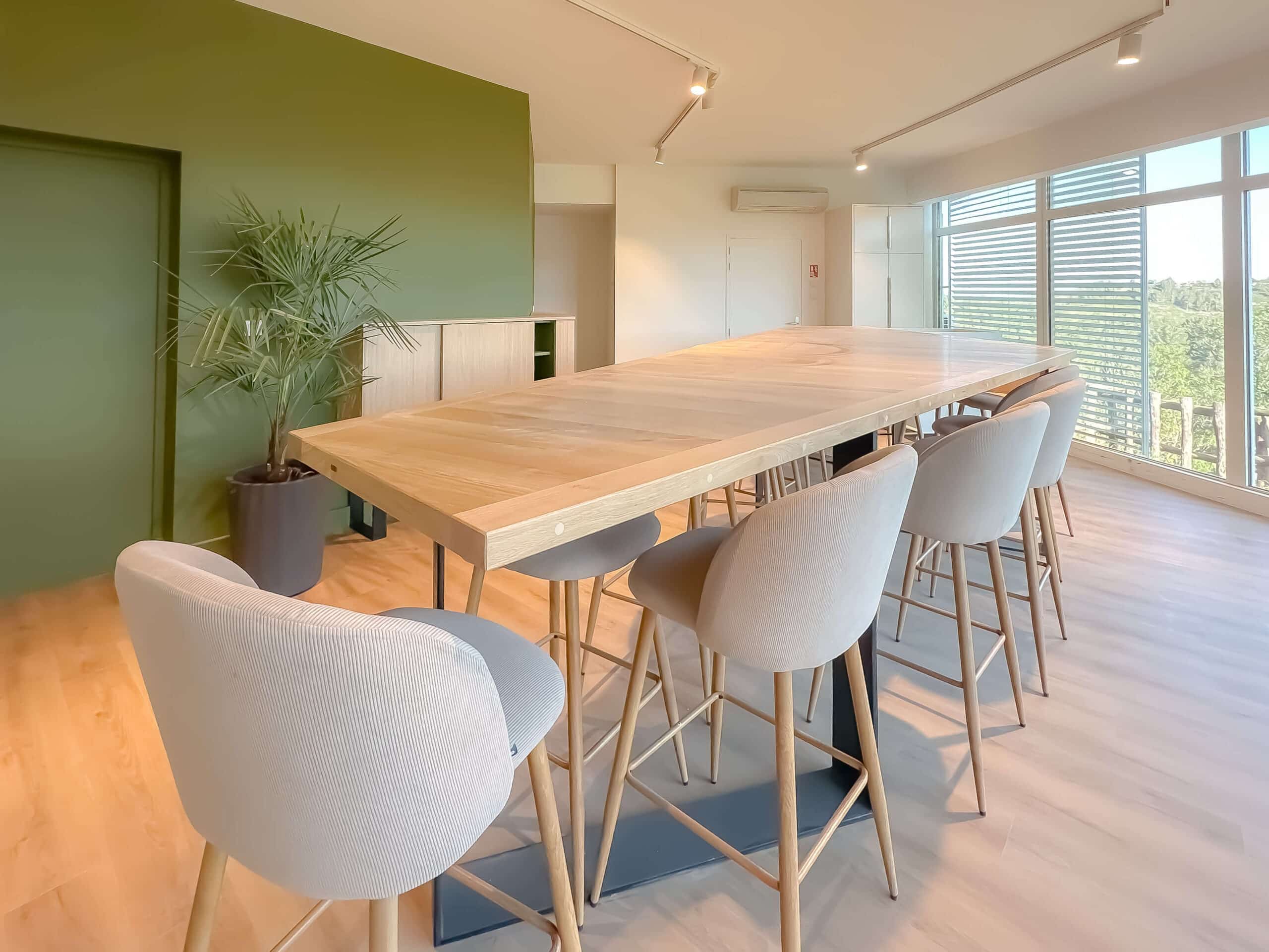 Table haute bois, tabourets gris, mur vert et plante dans salle de réunion moderne et lumineuse.