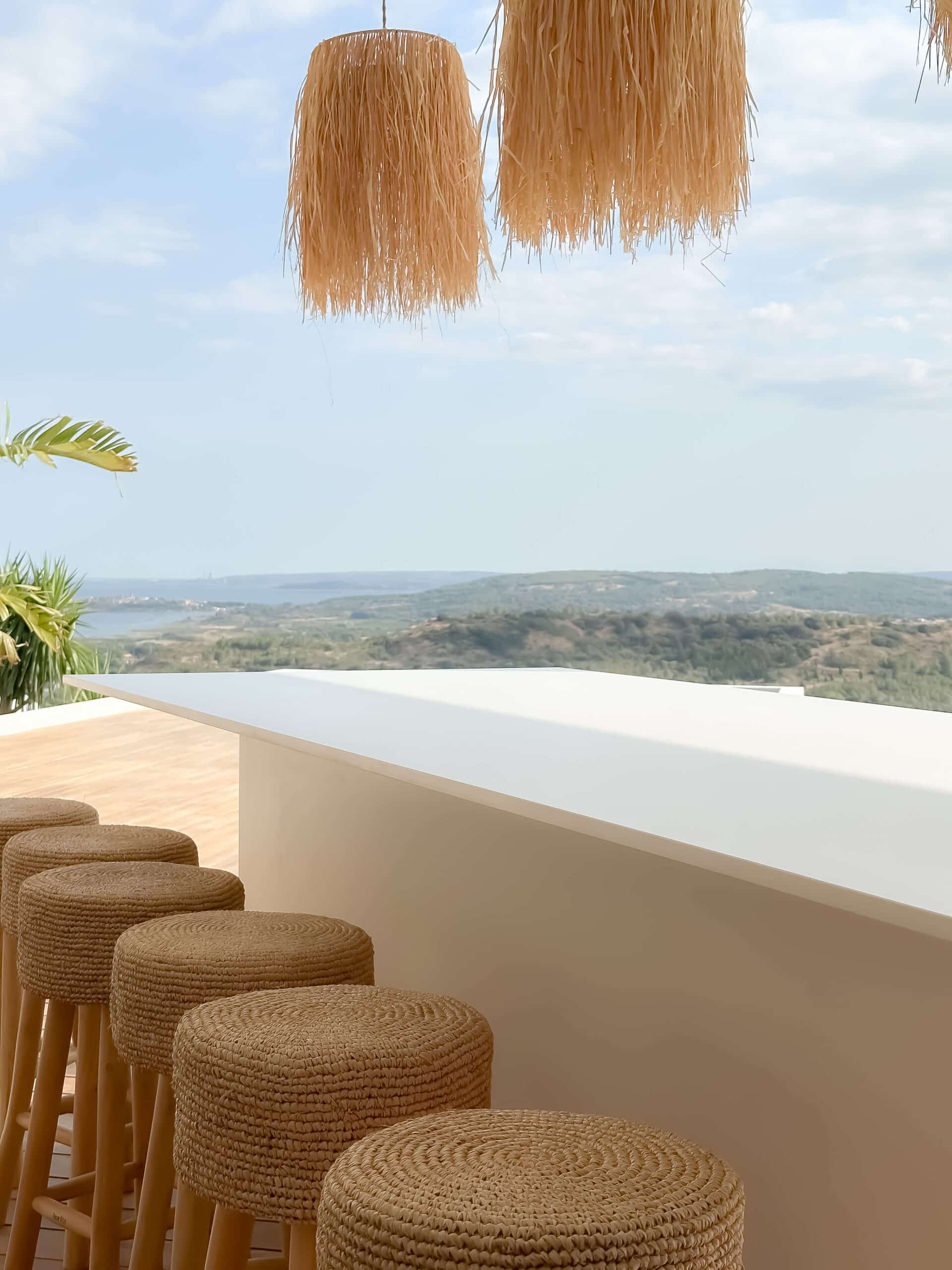 Bar de terrasse blanc, tabourets en paille et luminaires à franges avec vue sur les collines.