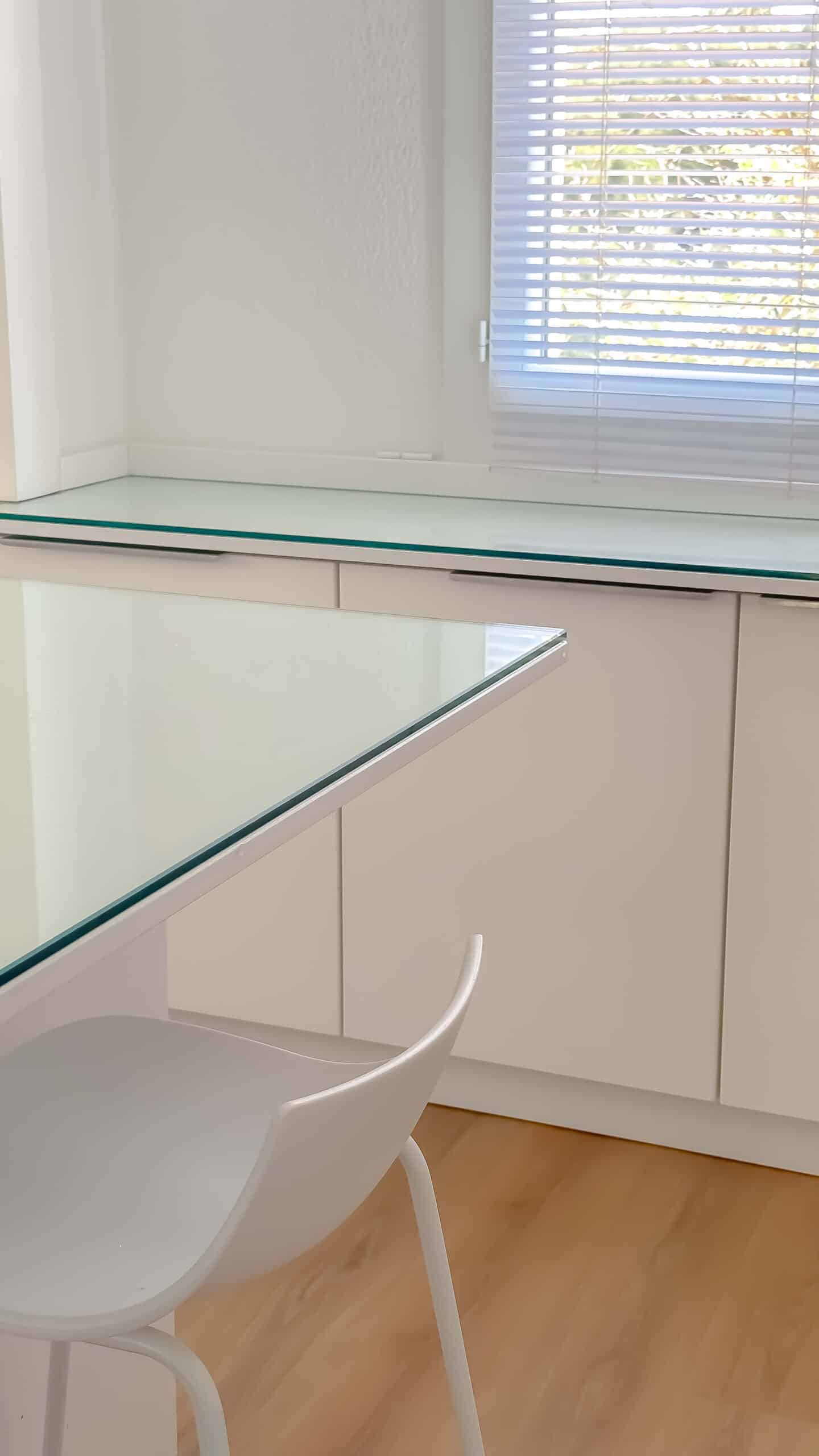 Cuisine blanche minimaliste avec table en verre, chaise design et meubles épurés sous une fenêtre à stores.