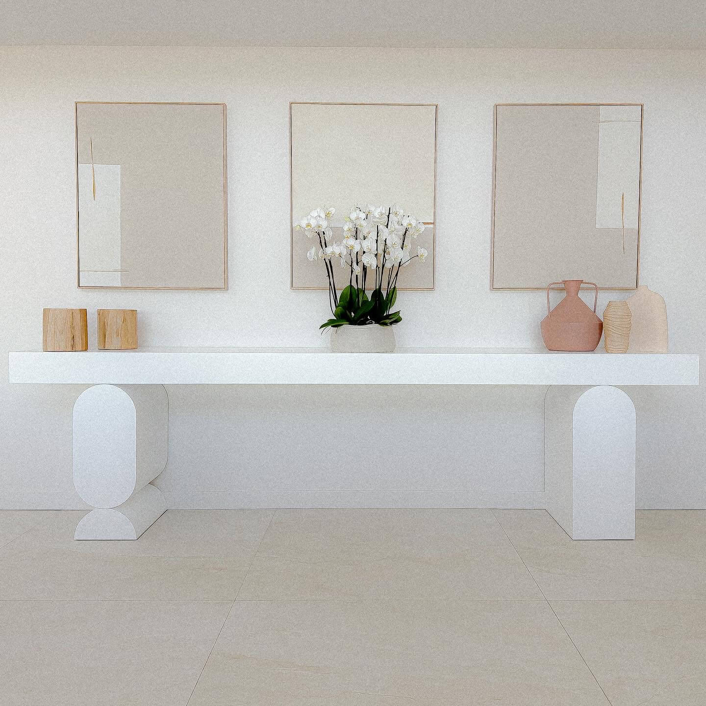 Console blanche, orchidée centrale, vases et trois cadres minimalistes sur mur clair.