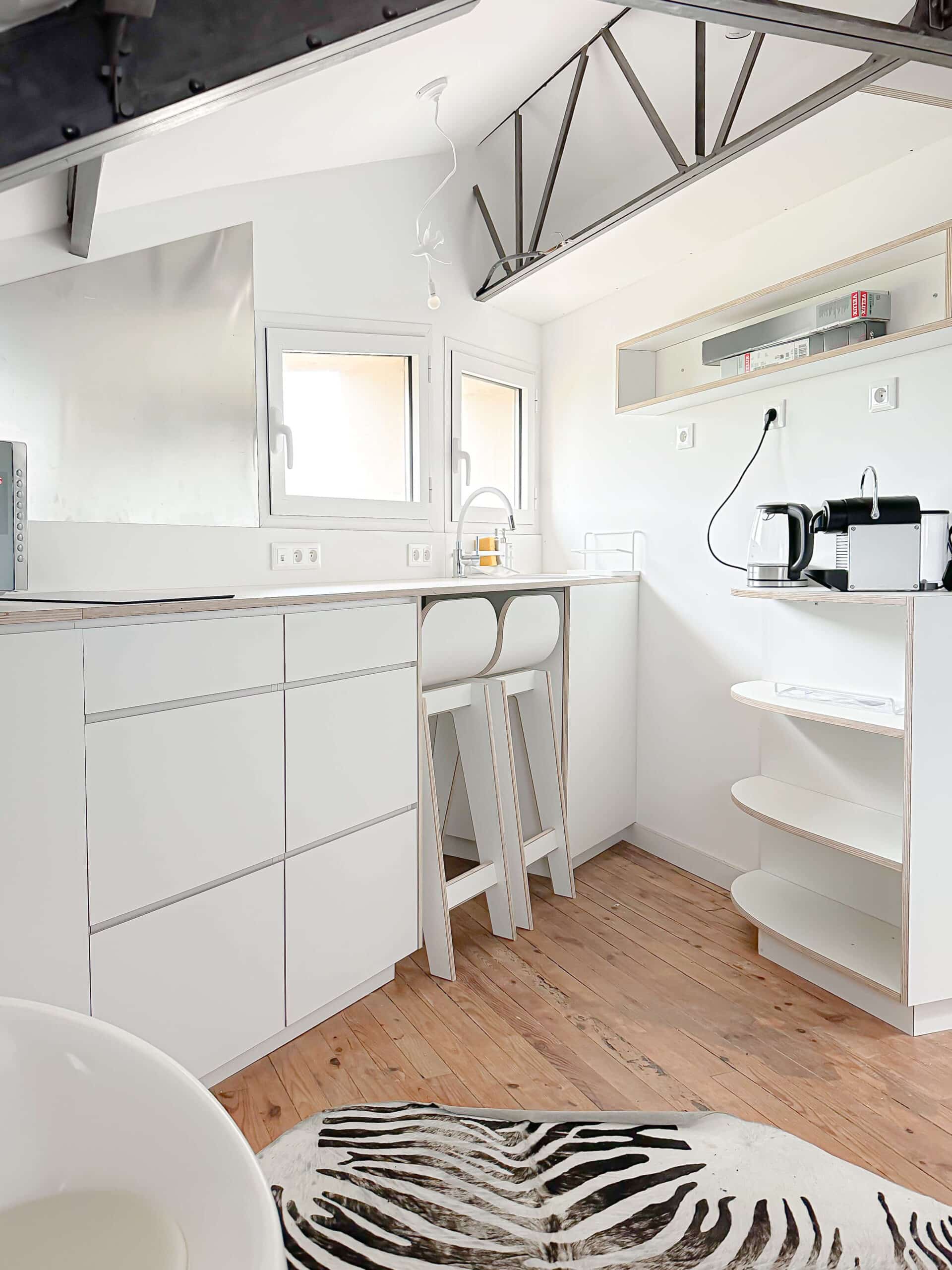 Cuisine moderne blanche minimaliste avec sol en bois, tabourets hauts et tapis zébré dans un loft lumineux.