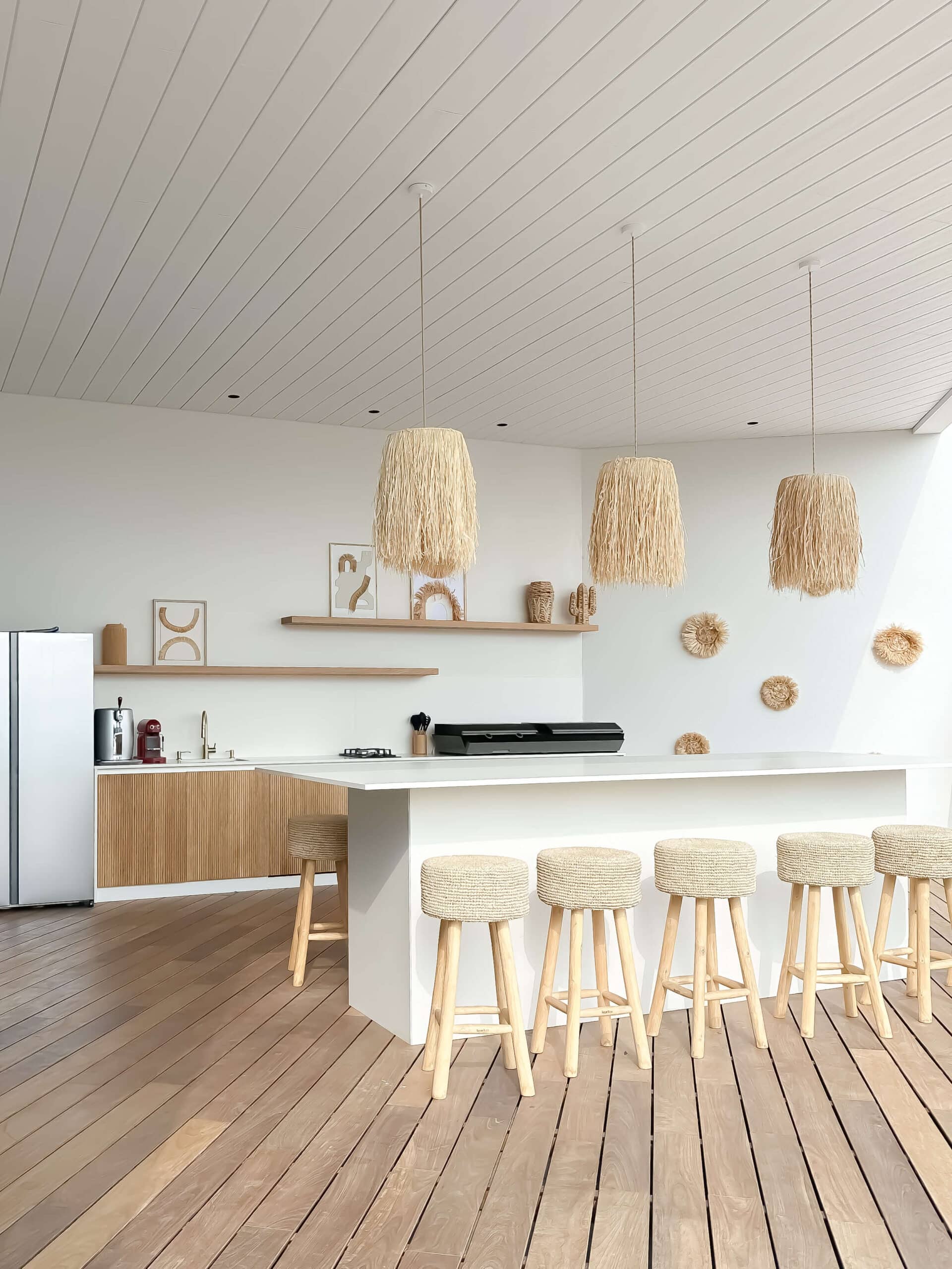 Cuisine d'été au style boho avec îlot blanc, tabourets tressés et suspensions en paille sur terrasse en bois.