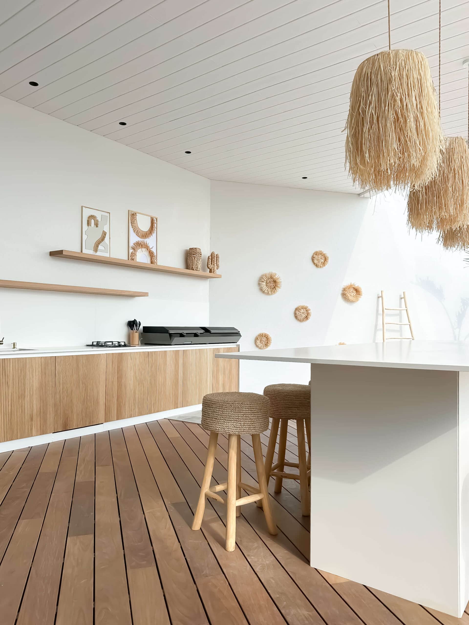 Cuisine extérieure moderne bohème avec îlot blanc, tabourets en jute, luminaires en paille et terrasse en bois.