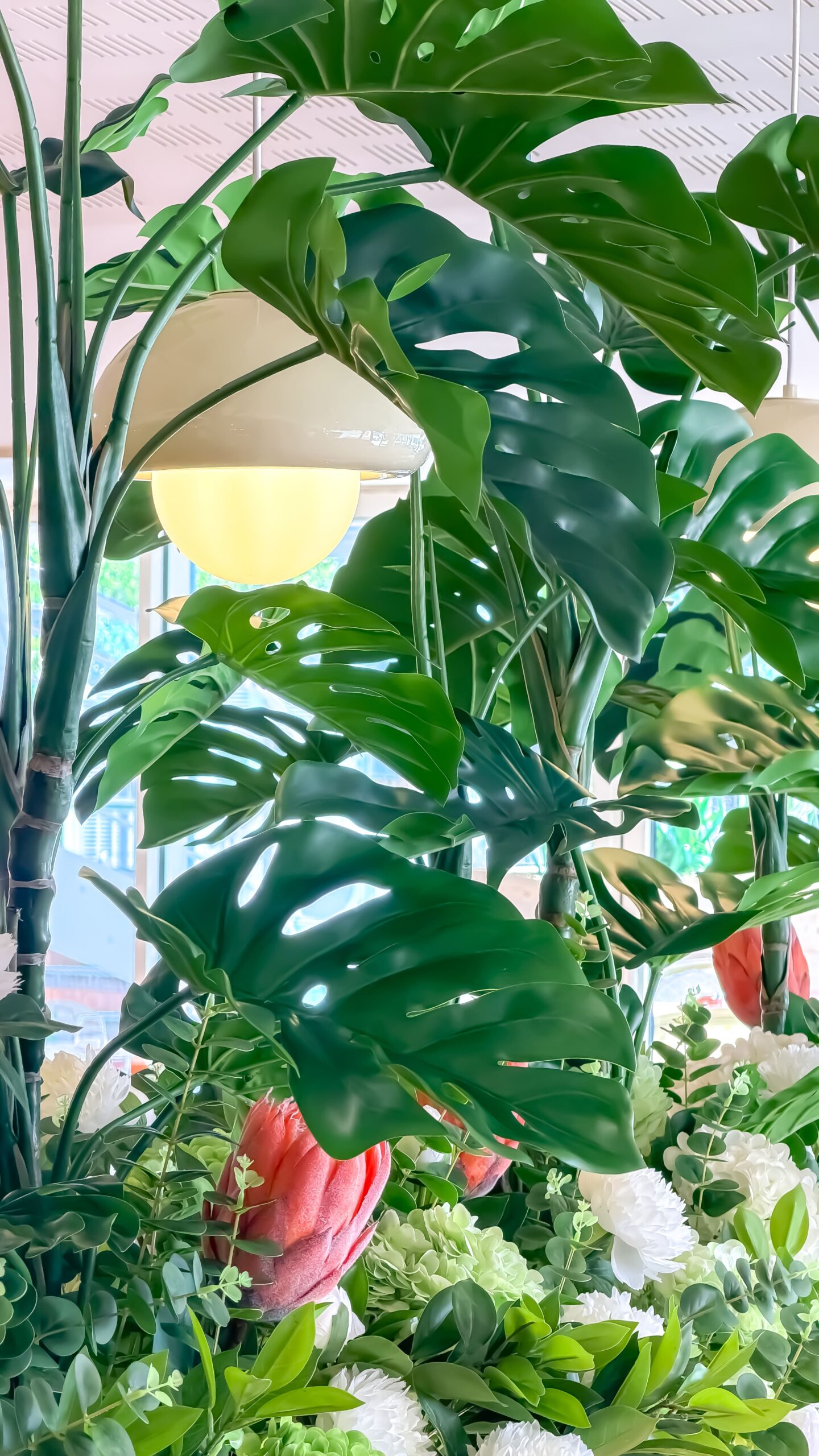 Grandes feuilles de monstera entourant une lampe design crème avec fleurs exotiques rouges et blanches en dessous.