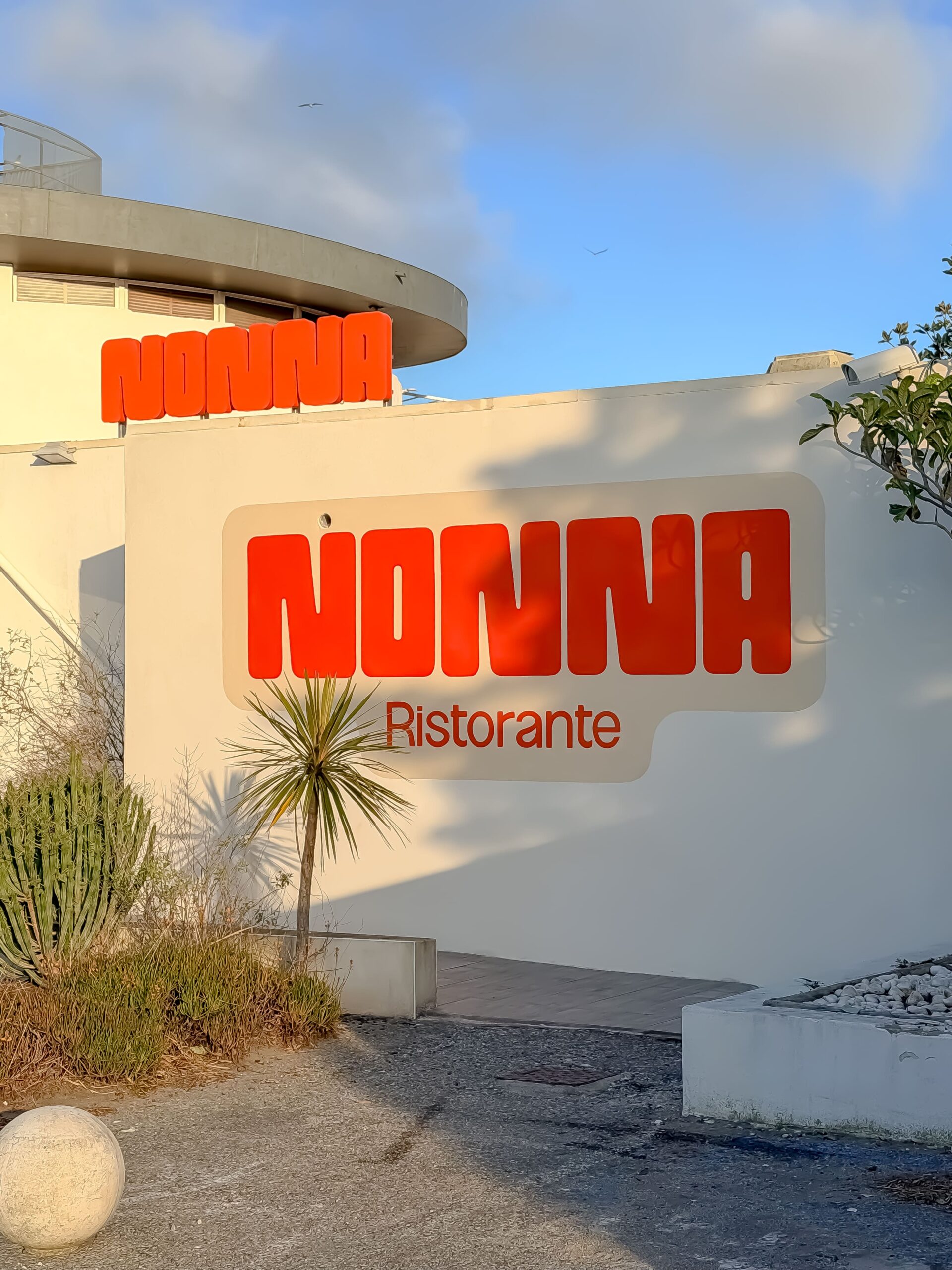 Enseigne orange NONNA Ristorante sur façade blanche avec palmier sous un ciel bleu ensoleillé.