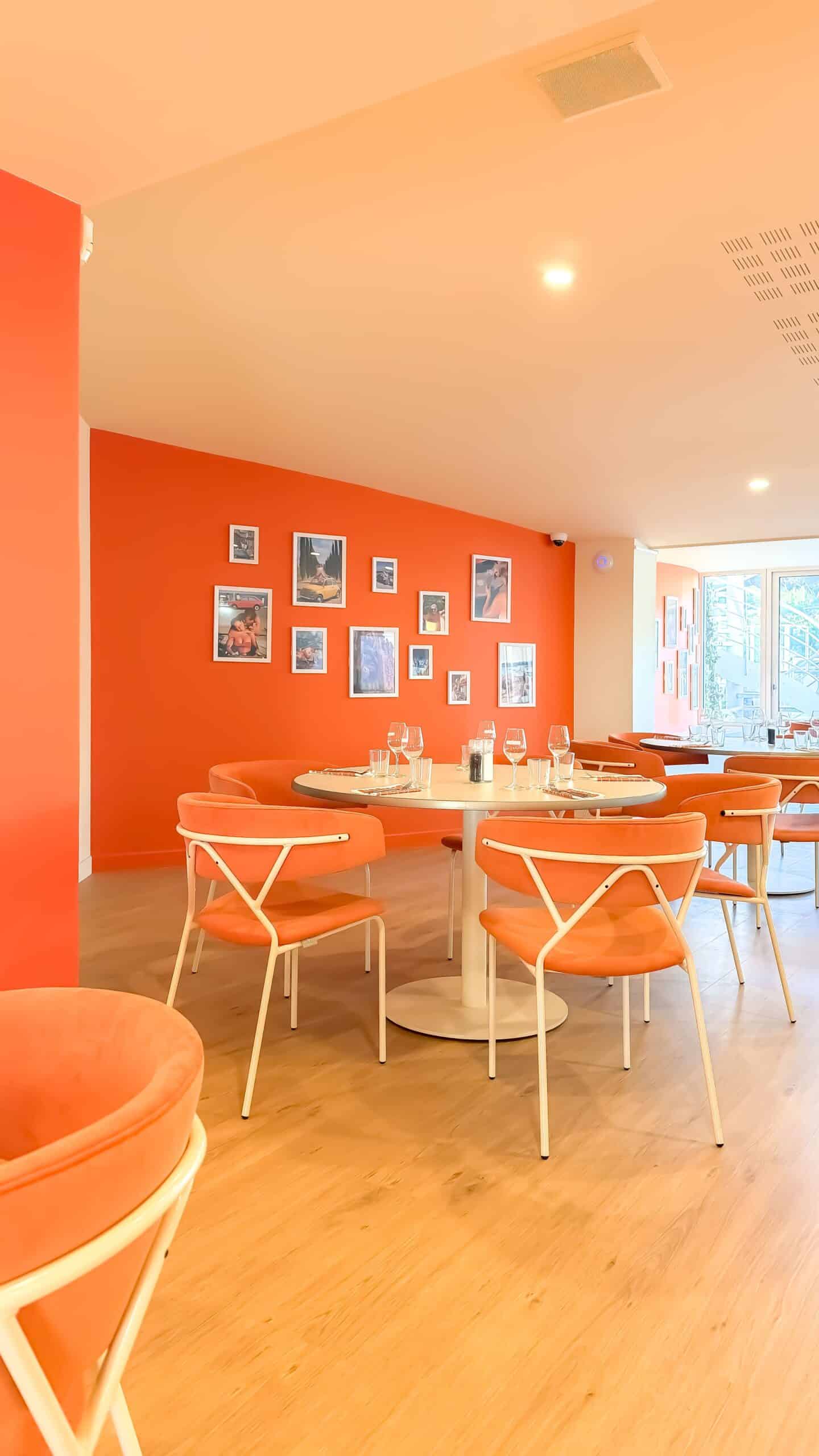 Restaurant au décor orange moderne avec chaises design, table ronde dressée et galerie de photos au mur.