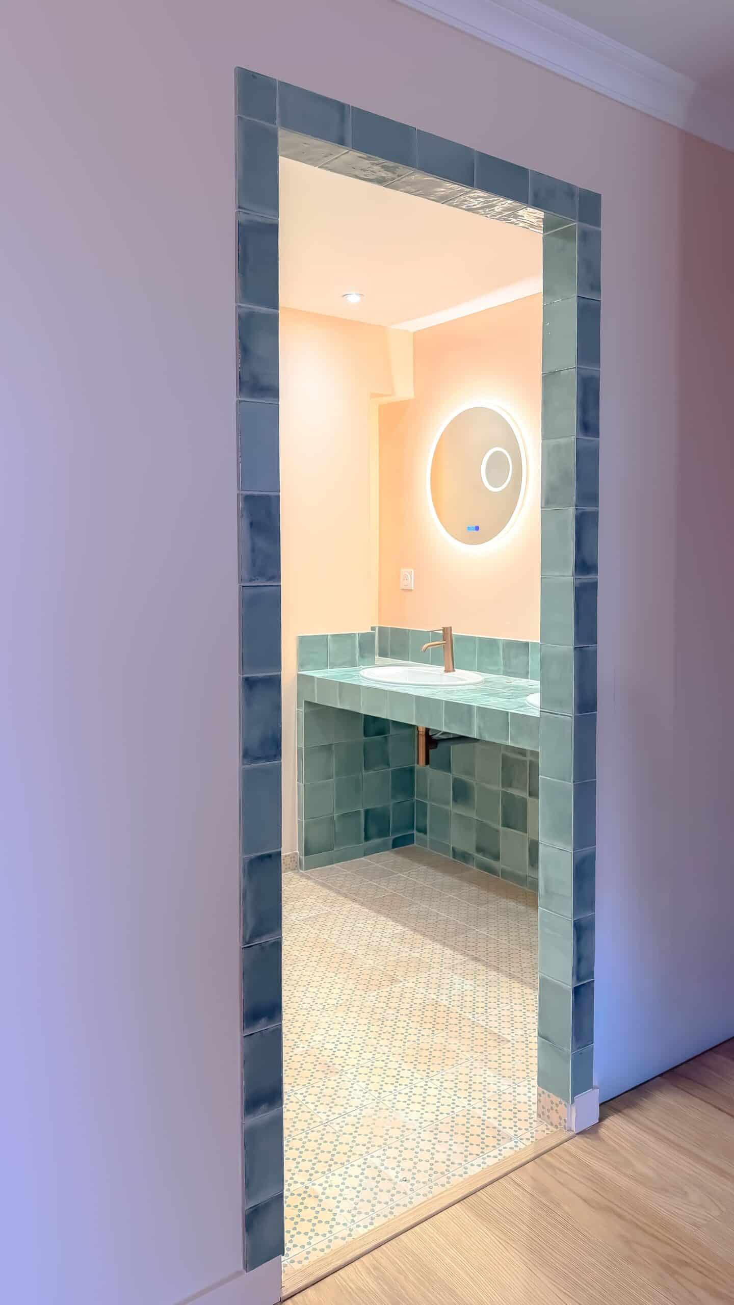 Salle de bain avec carrelage bleu canard, murs pêche, miroir rond lumineux et robinetterie dorée.