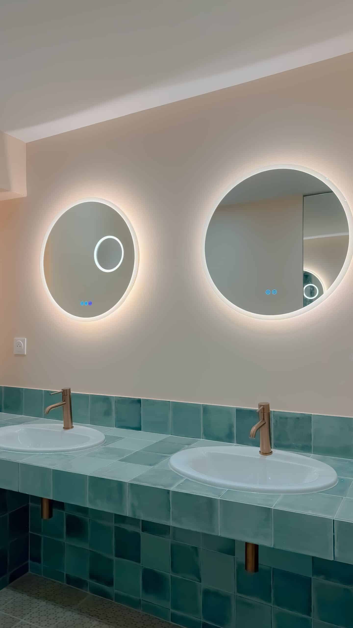 Double vasque, carrelage bleu, miroirs ronds rétroéclairés et robinets dorés élégants.