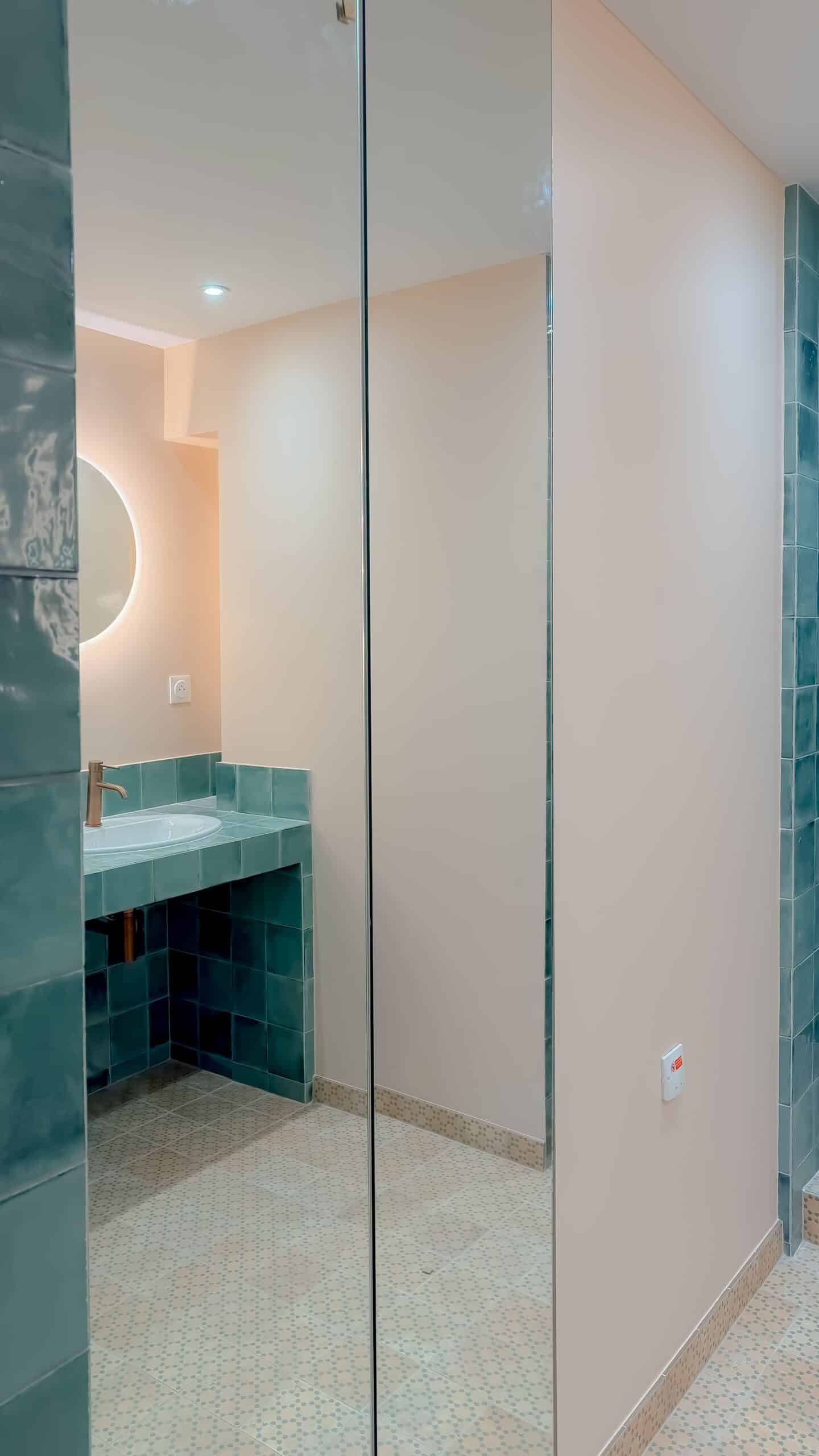 Salle de bain moderne : carrelage bleu canard, robinet doré et grand miroir sur mur crème épuré.
