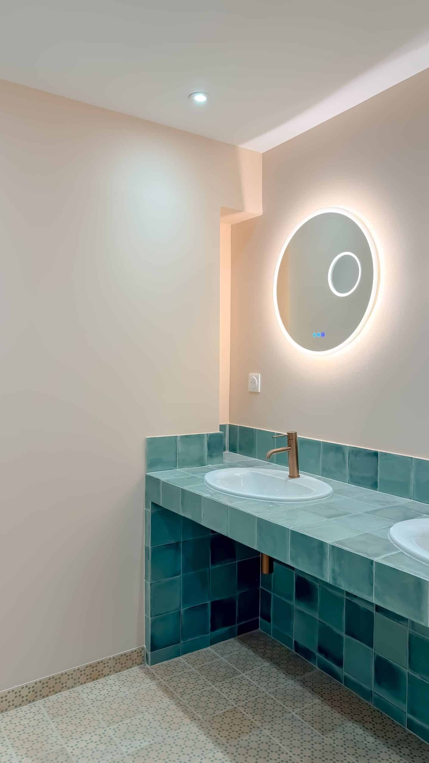 Salle de bain : vanité en carreaux turquoise, robinet or et miroir rond lumineux sur mur beige.