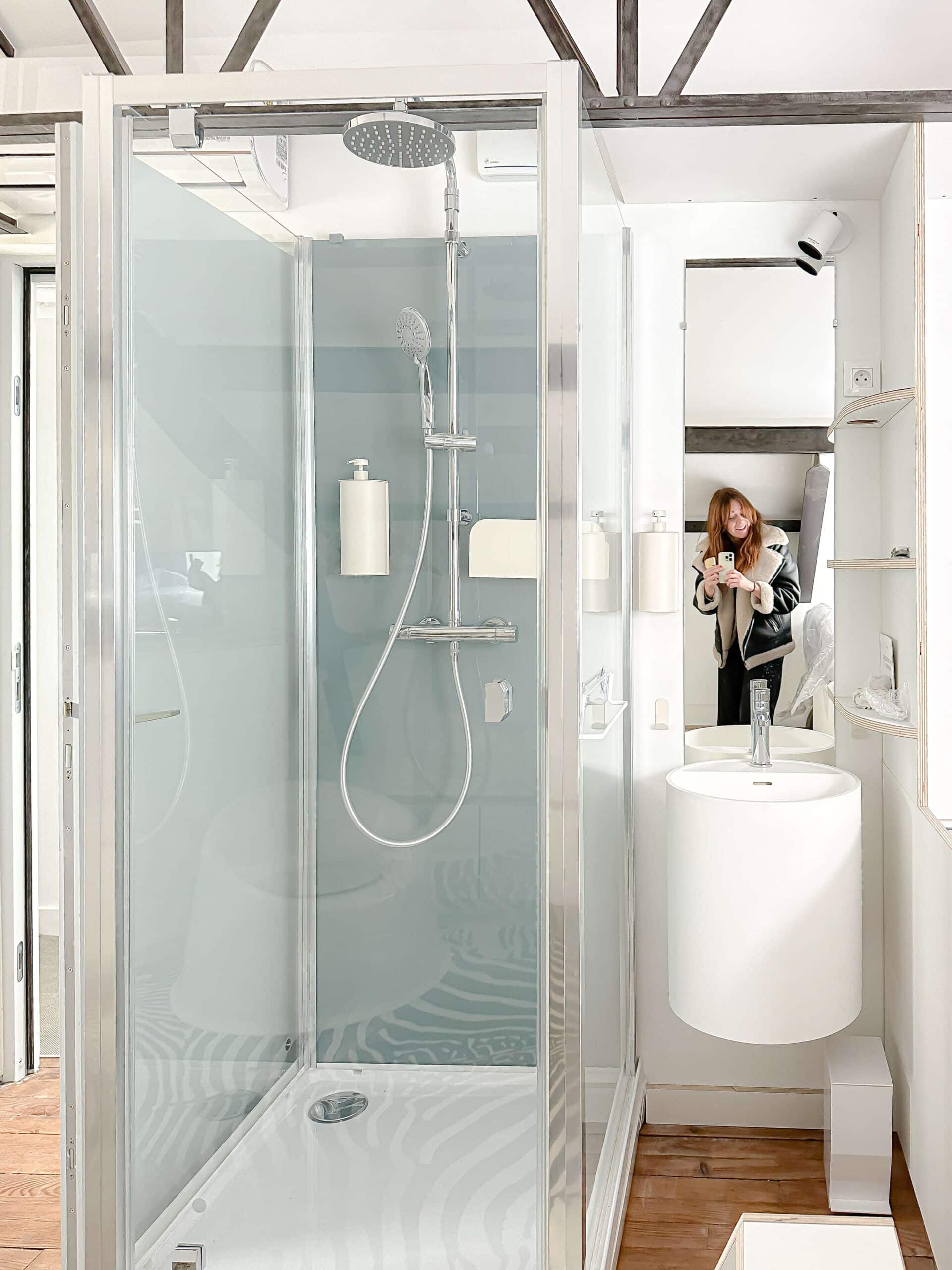 Douche design vitrée, vasque blanche et miroir reflétant une femme souriante dans une salle de bain moderne.